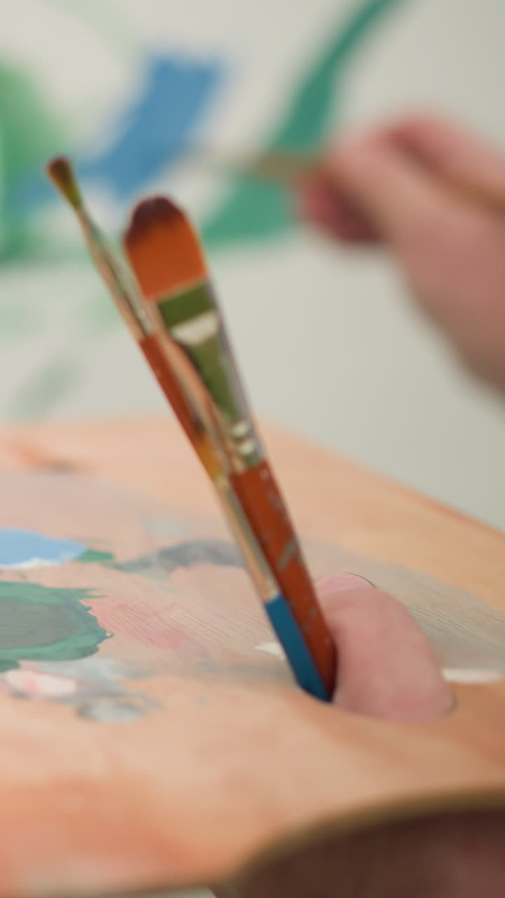 se ve a un pintor, cuya cara no es visible, sosteniendo una paleta de madera llena de varios colores de pintura vibrantes. vestido con una camisa a cuadros, el artista se centra en aplicar pintura a un lienzo grande