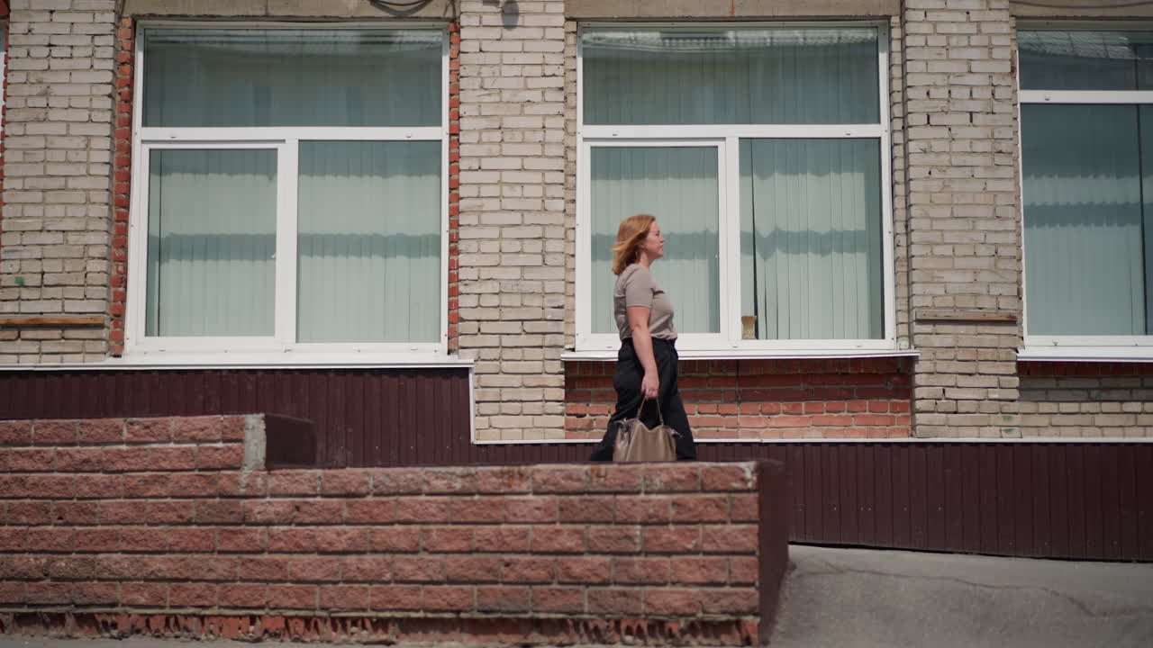 Side view of pupil walking out of building carrying handbag under bright sunlight, brick wall and large windows reflecting calm academic environment