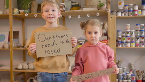 niño rubio y niña rubia en un taller de artesanía sosteniendo dos carteles de cartón con frases de que no hay planeta b y nuestro planeta necesita ser amado