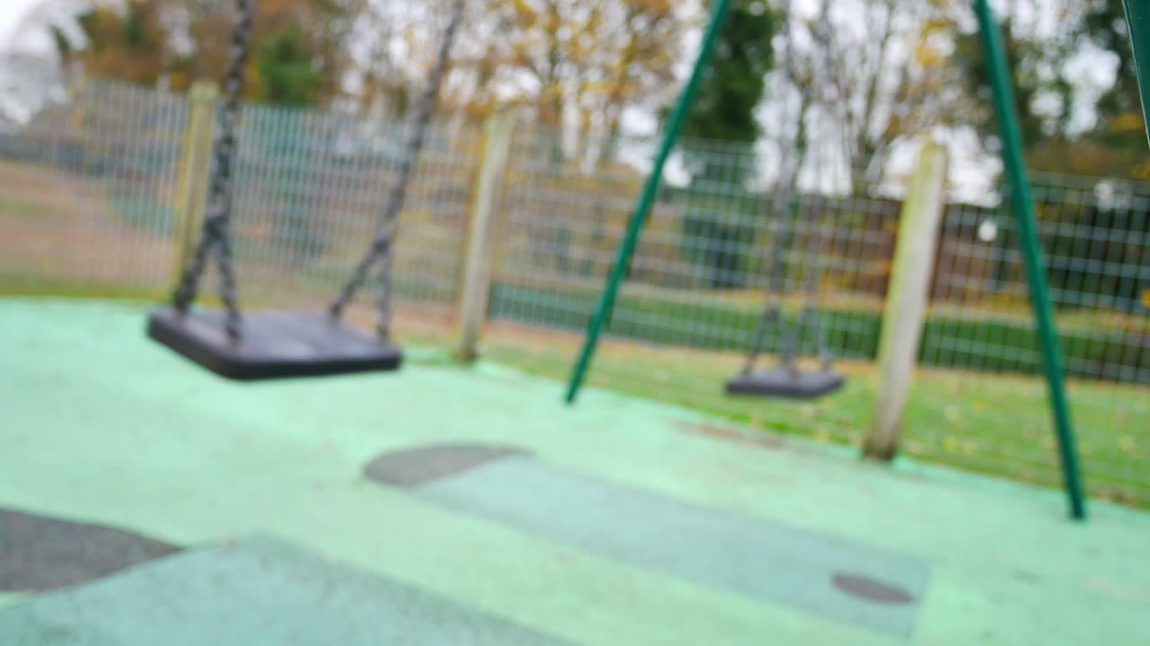 Emerging shot from behind bar to view unfocused swings swinging in empty playground
