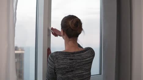 back view of young woman opening her bedroom curtains and the window and looking outside in slow motion