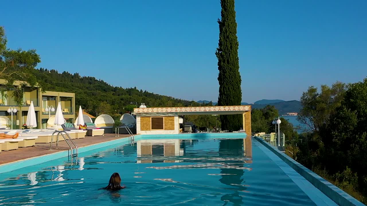 Tracking shot of hotel terrace with sun beds. Young woman swims through the pool.