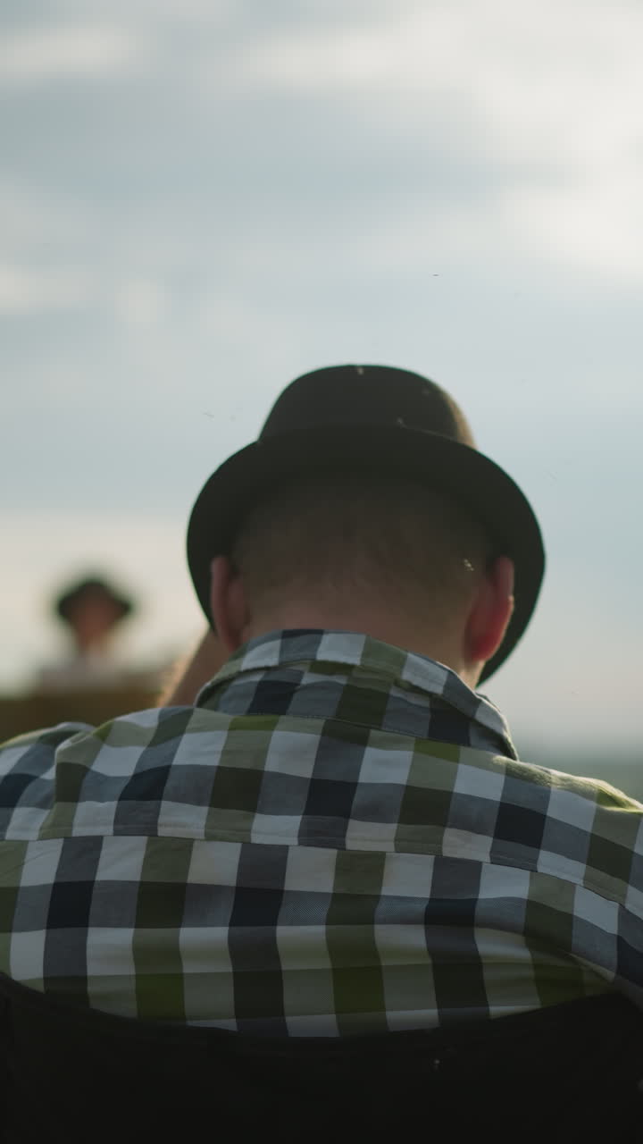 검은 모자를 입은 남자가 잔디에 앉아있는 동안 그것을 조정하는 근접적인 뒷면. 모기는 배경에 흐릿한 여자가 그림을 그리면서 그의 주위에 날아갑니다.