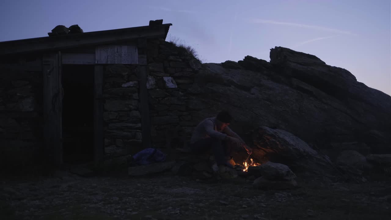 A person tends a campfire outside a stone hut at dusk in the mountains.