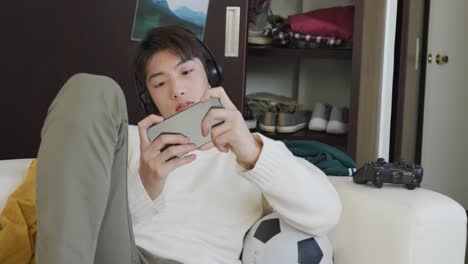 Asian-boy-wearing-headphones-playing-game-on-smartphone-sitting-on-the-couch-at-home