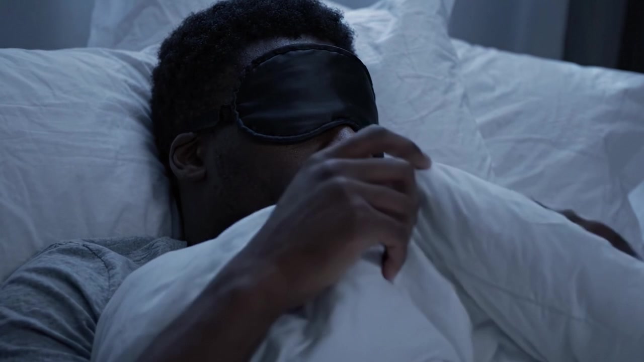 En ung mand med sovemaske hviler komfortabelt i sengen omgivet af bløde puder under en hyggelig dyne, der omfavner ro og fredelig søvn.