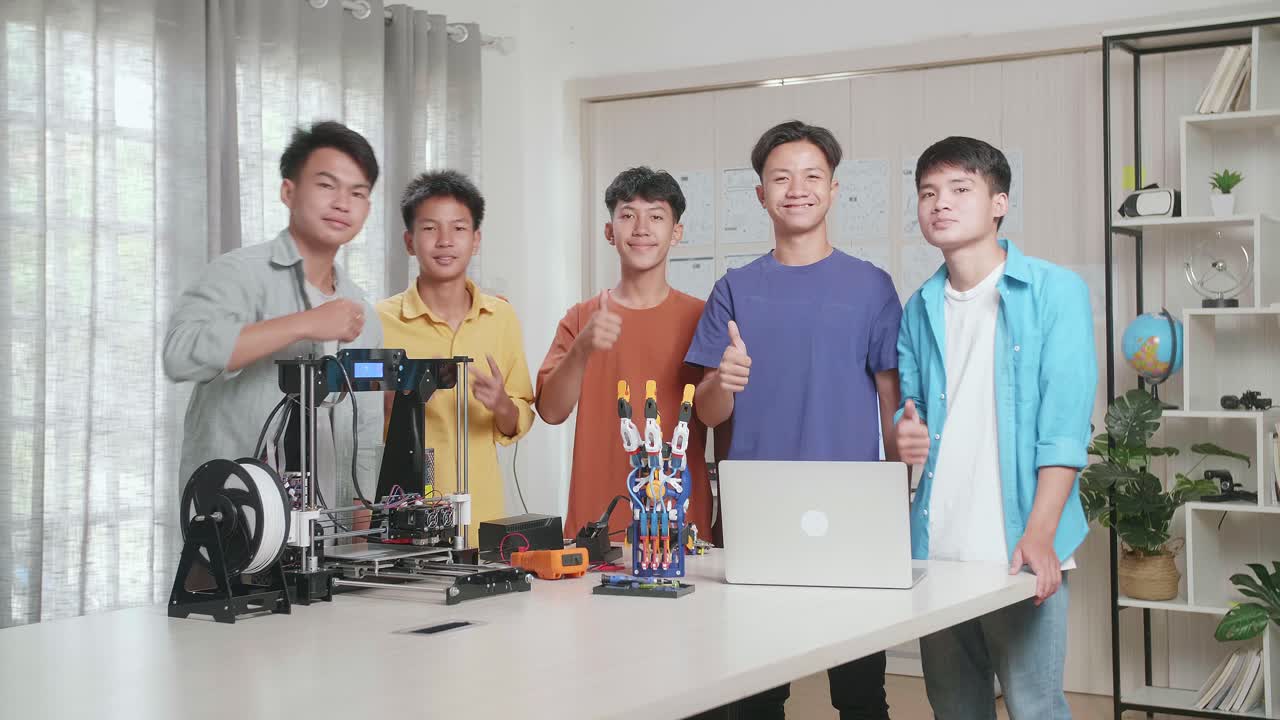 노트북과 3d 프린팅을 가진 젊은 아시아 소년들의 그룹은 집에서 사이보그 손에 대해 논의하는 동안 미소 짓고 엄지손가락을 카메라로 제스처를 보여줍니다.