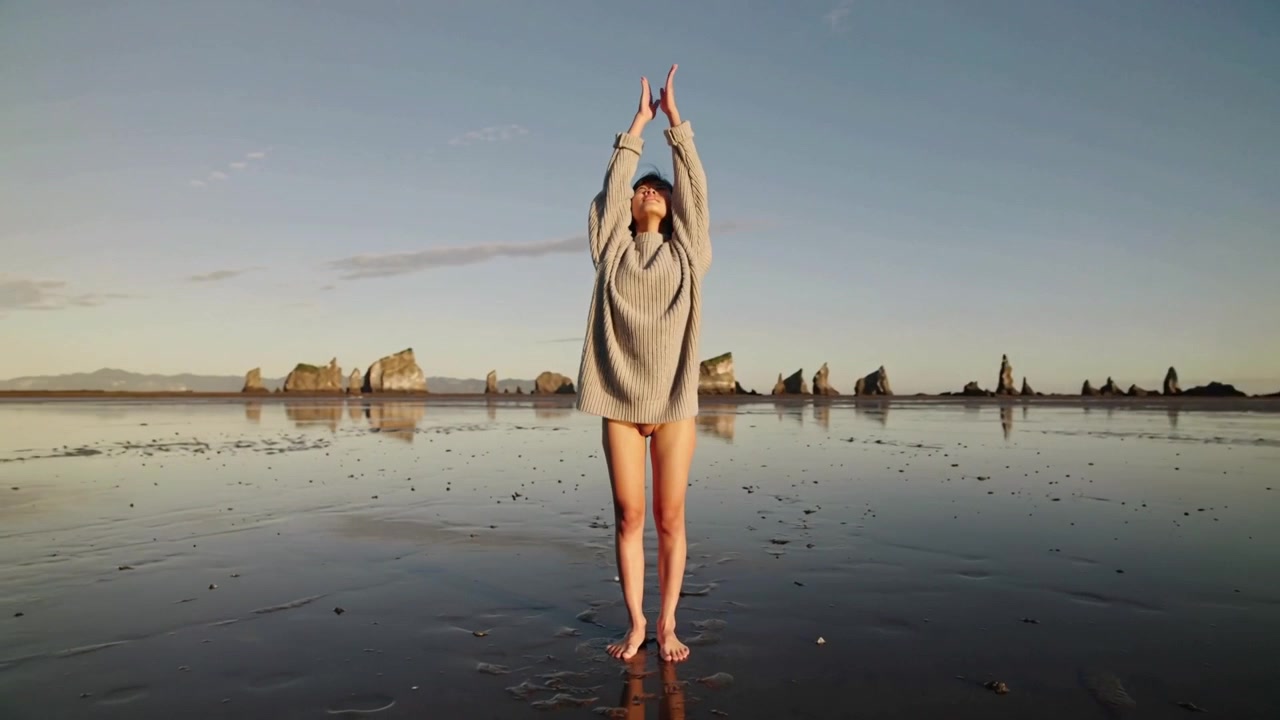 Una donna che si allunga con un maglione oversize, sollevando i palmi in alto su una spiaggia con la marea, saluta i faraglioni all'alba.