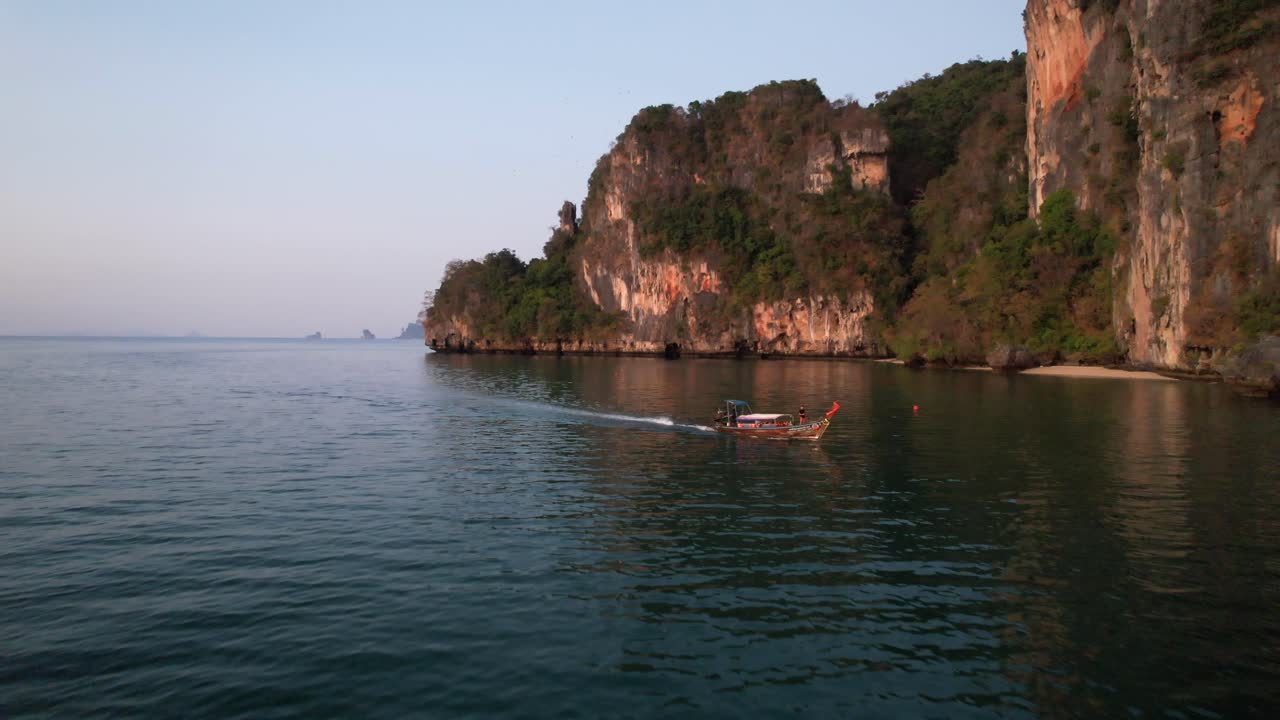 Ripresa aerea con drone che si allontana vicino a Railay Beach a Krabi, Thailandia, durante l'alba, mentre una barca longtail thailandese naviga nel Mare delle Andamane con un turista a bordo e grandi montagne calcaree sullo sfondo
