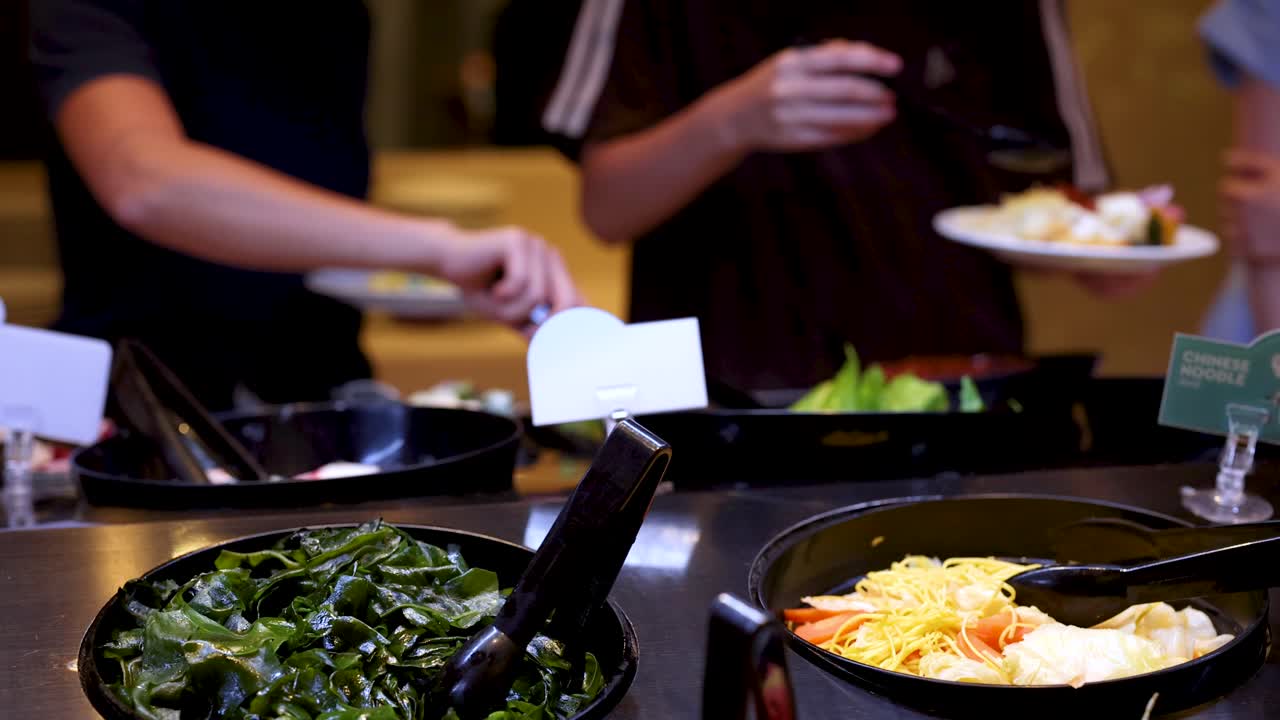 Diners select fresh salad greens with tongs at buffet-style restaurant under warm indoor lighting