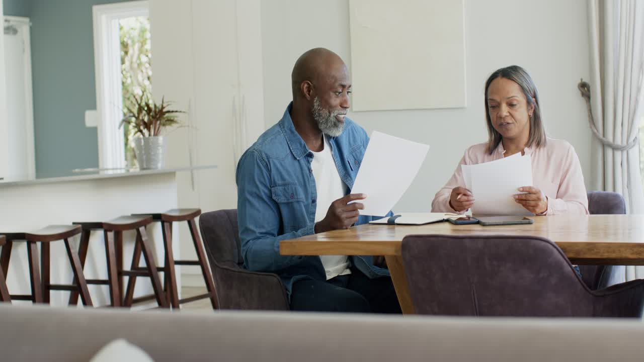 Senior couple sitting at dining table, man holding sheet initiating sorting bills together