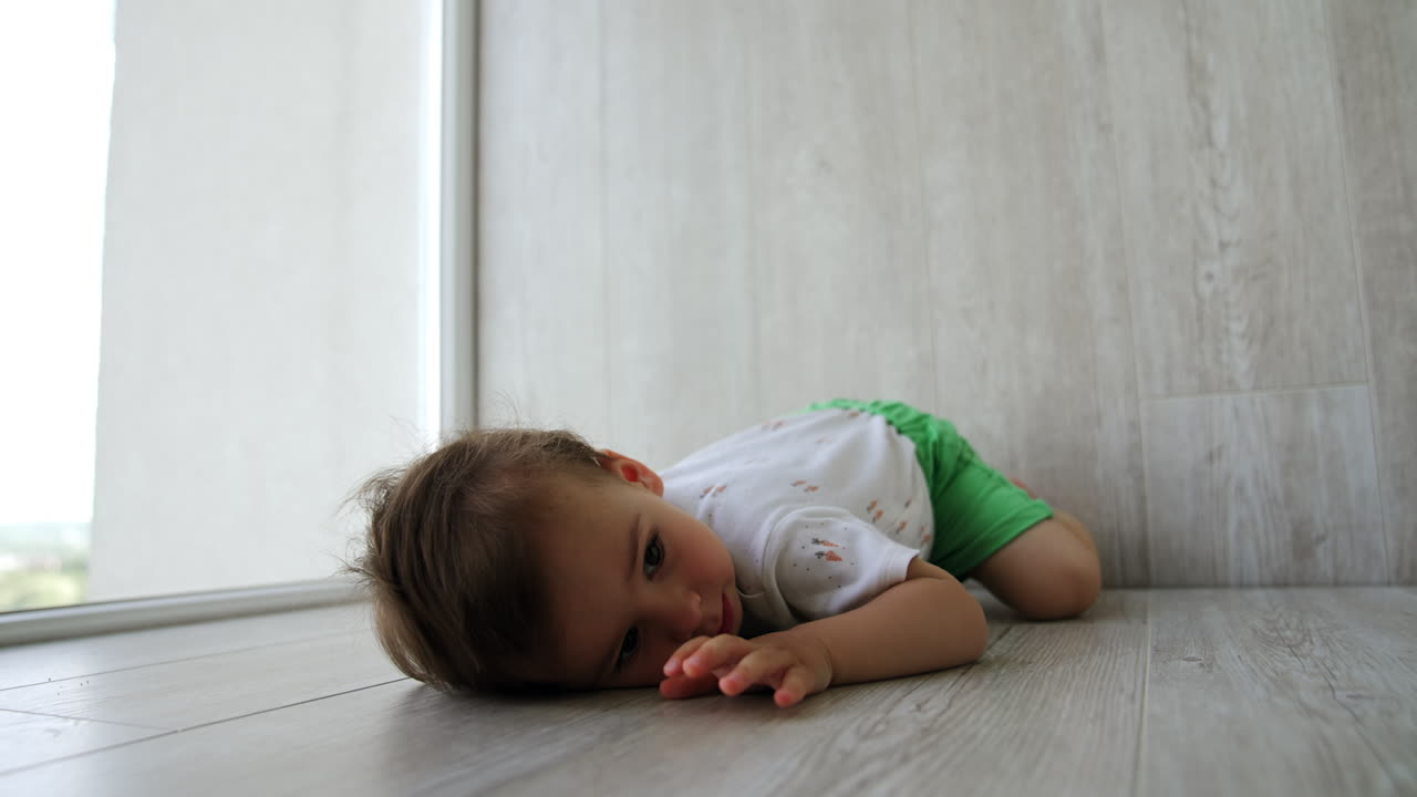 Adorable baby boy plays at home. Kid sits on the floor, puts his cheek to the floor and stands up again.