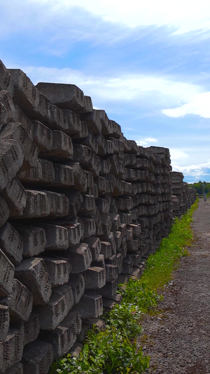 Grande pila di vecchie e logore traversine ferroviarie in cemento impilate per il riciclo o il riutilizzo in un sito industriale accanto a un binario ferroviario sotto un cielo blu con alcune nuvole
