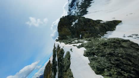 Aerial-FPV-view-of-a-snowy-mountain-slope-with-patches-of-exposed-rock-and-greenery,-framed-by-distant-mountain-peaks-and-a-lush-valley-below,-representing-the-beauty-of-nature