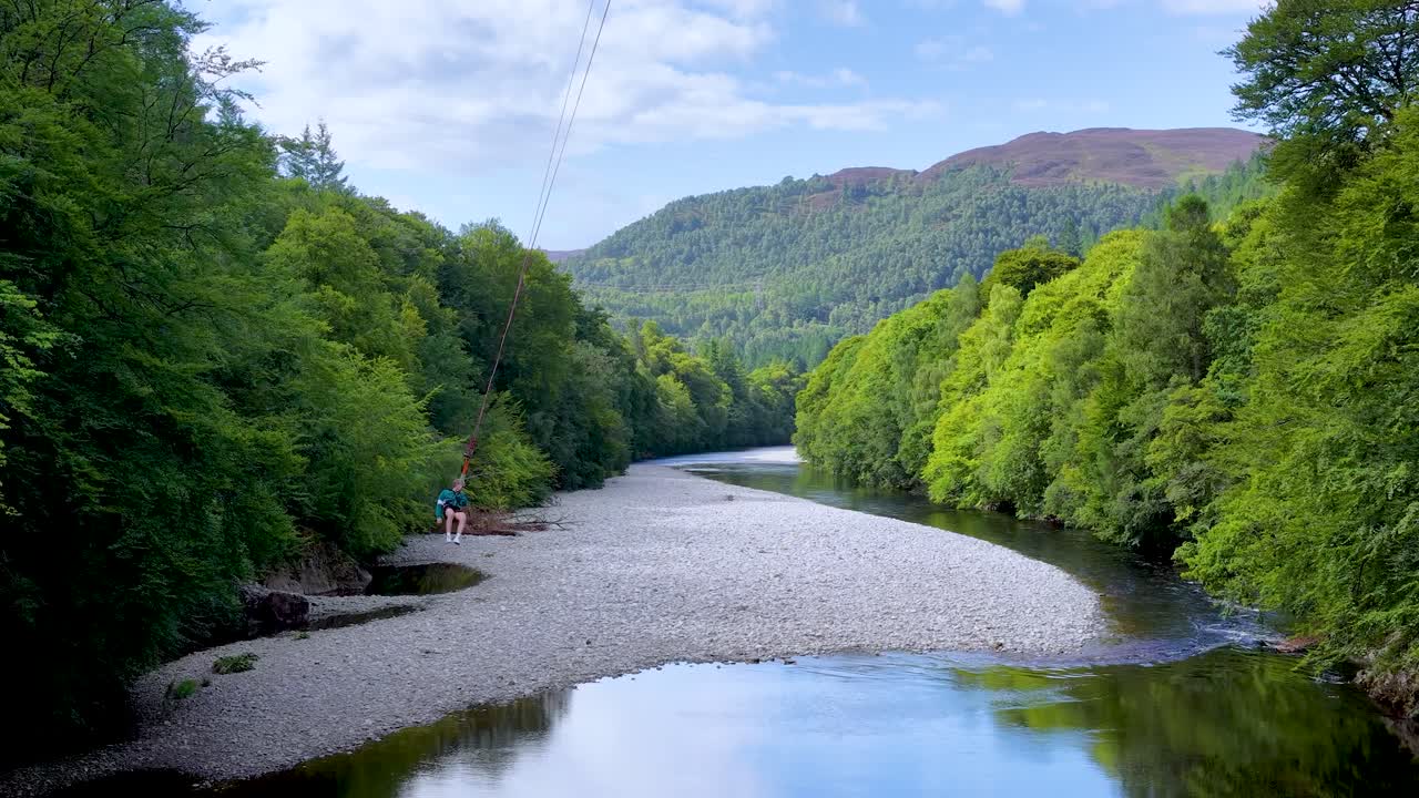 Individual rides zipline above river, lush green forest, bright daylight, wide static camera shot