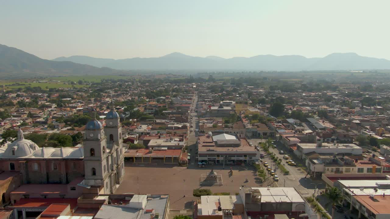 Wide aerial view of Tuxpan Jalisco with church, plaza, city market and distant mountains. Slow dolly in droneshot