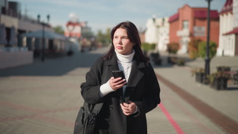 stylish woman in black coat and white sweater walks on sunny urban street, focused on her smartphone, background features charming architecture, blurred greenery, and paved pedestrian pathway