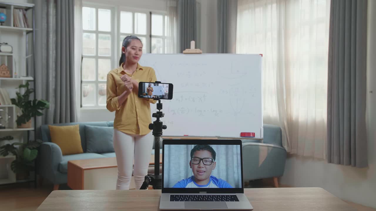 Asian Female Teacher Shoots Video By Smartphone Camera And Having Video Call On Laptop While Teaching Math At Home