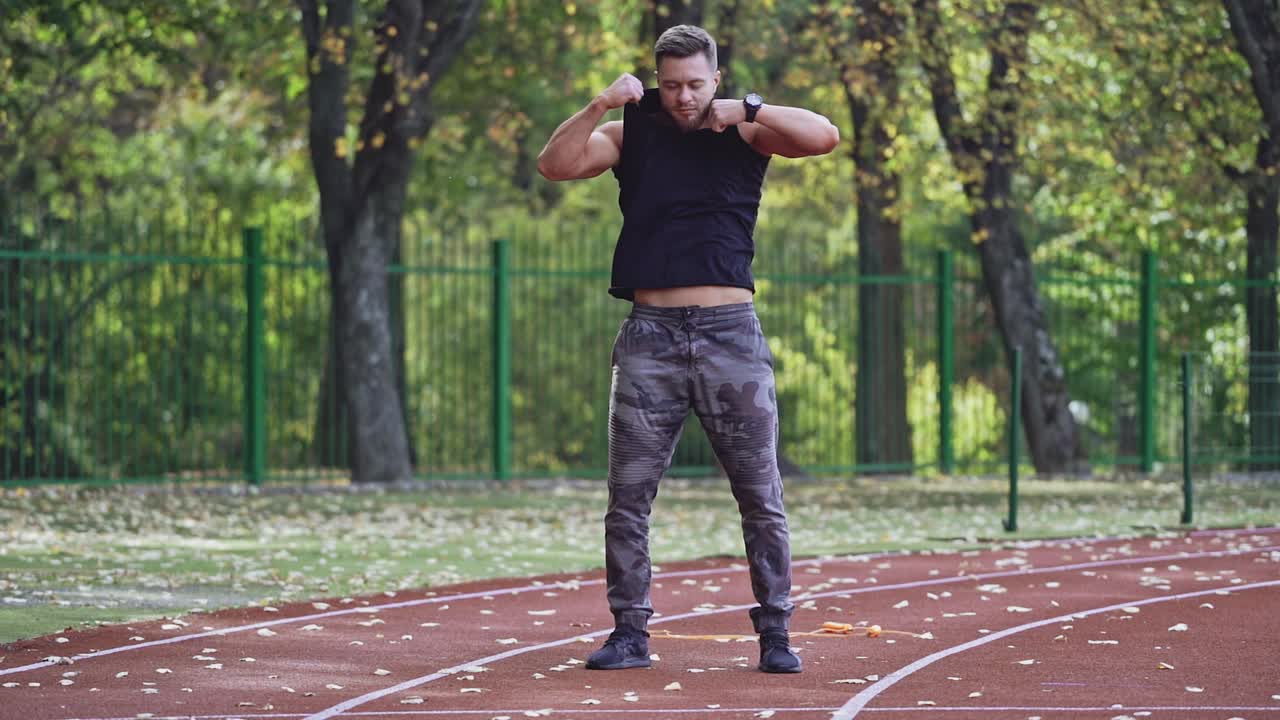 Full-length portrait of athlete undressing outdoors. Handsome man taking off his black shirt while standing on the stadium. Slow motion.