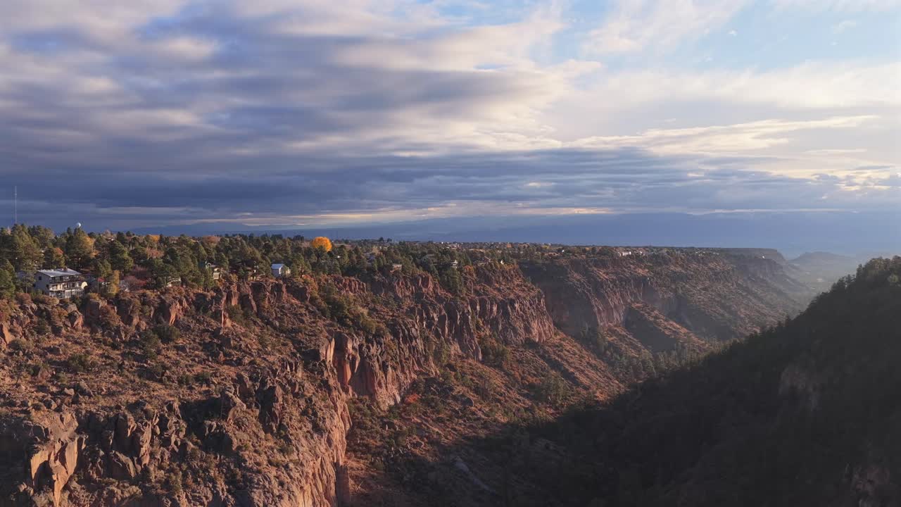 Dynamic aerial rise-and-pan shot revealing a sprawling southwest town built along the canyon rim. The vast gorge contrasts sharply with the nearby residential architecture at sunrise