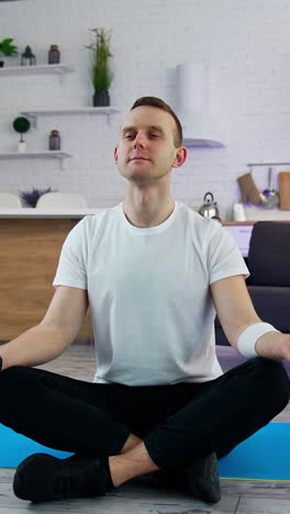 young man sitting in yoga pose in the kitchen. guy in white t-shirt doing relax exercises after workout while sitting on a mat at home.. vertical video
