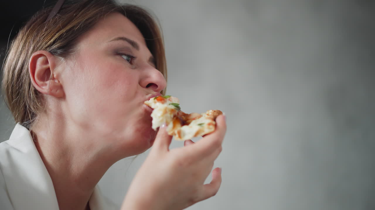Close up of lady in white outfit eating pizza slice with melted cheese and toppings, enjoying every bite with playful expression, holding pizza near mouth against cozy blurred background