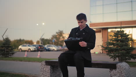 business mogul in black suit walks up to bench, opens briefcase in outdoor setting with modern building and parked cars in background, professional, urban office worker taking a break