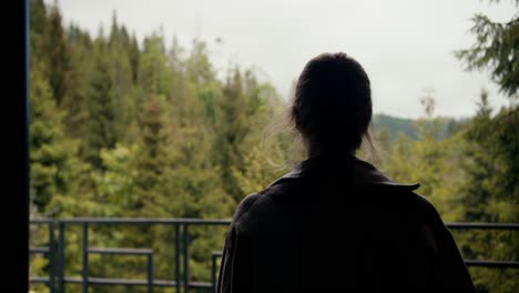 a happy blonde girl with a cup of coffee goes to the balcony of a country house and looks at the lush green coniferous forest in the mountains