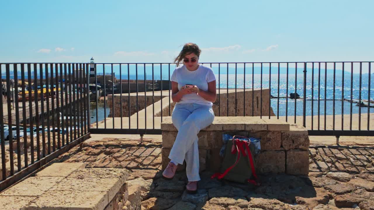 Turista seduto su una panchina che usa lo smartphone nell'antico porto di Akko.