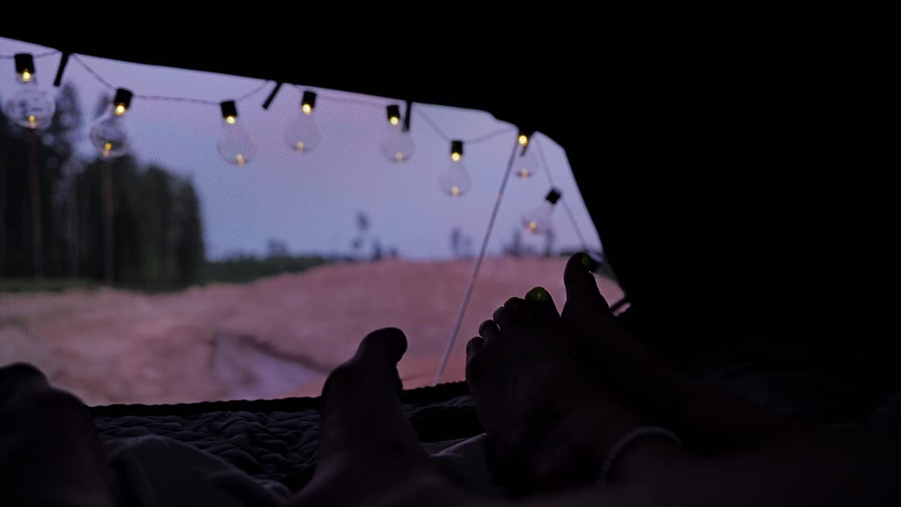 Feet of couple rest inside rooftop tent at dusk with glowing string lights