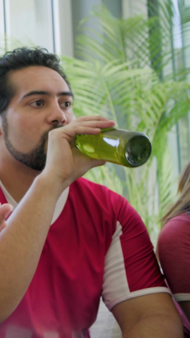 Vertical video: Reacting to offscreen score man in red tee drinking green bottle on couch by window