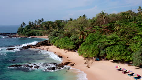 slow drone push-in with a gentle pan, starting from colorful beach umbrellas and revealing a tropical palm shoreline and turquoise sea at silent beach, sri lanka.
