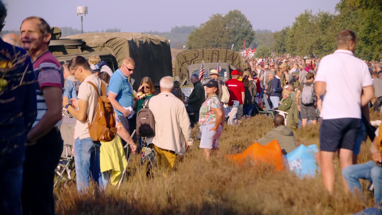 Crowd gathered at Airborne Ede event, watching a mass parachute drop on a sunny day. Families, individuals, and military enthusiasts stand or sit in the grass, enjoying the historical reenactment.