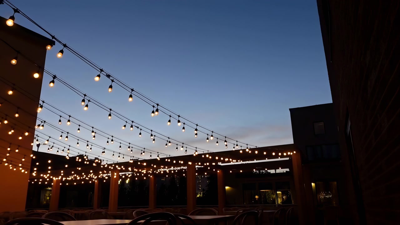 Warm hanging lights stretch across the top of a dark urban courtyard under a clear blue twilight sky, with building silhouettes and a few glowing background