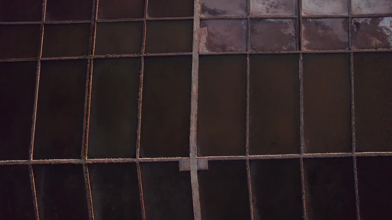 Geometric grid of dark brine evaporation ponds and narrow dikes forms the Salinas de Aveiro salt pans inside Ria de Aveiro coastal lagoon in Portugal, top down drone reveal shot
