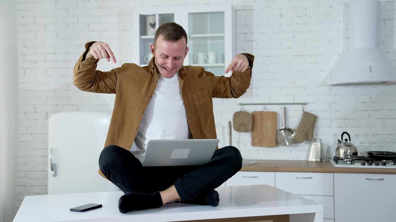 Funny man with a laptop on a table.Smiling freelancer sitting on a table and waiting to press button on a laptop on the kitchen background.