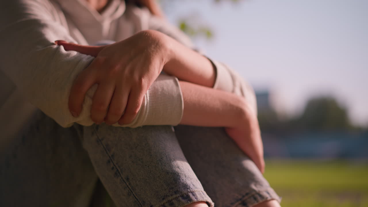close-up das mãos de uma jovem mulher descansando em joelhos dobrados em um ambiente ao ar livre pacífico, a luz do sol destacando seu aperto relaxado enquanto ela senta pensativa