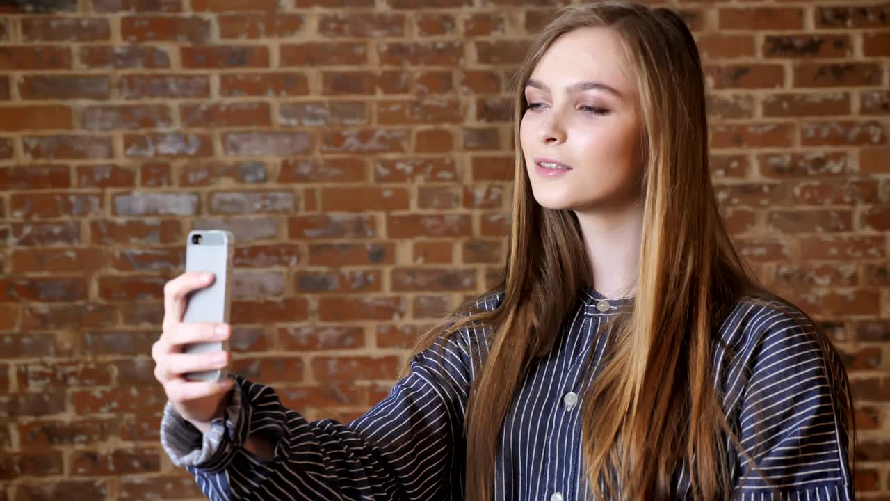 joven hermosa está haciendo selfie en su teléfono inteligente, sonriendo, tocando el cabello, concepto de comunicación, fondo de ladrillo