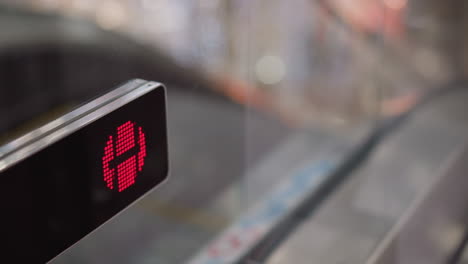 close up red warning indicator blinking on moving walkway glass barrier signaling stop, illuminated by soft bokeh mall lights, blurred escalator steps and shoppers moving behind transparent panel