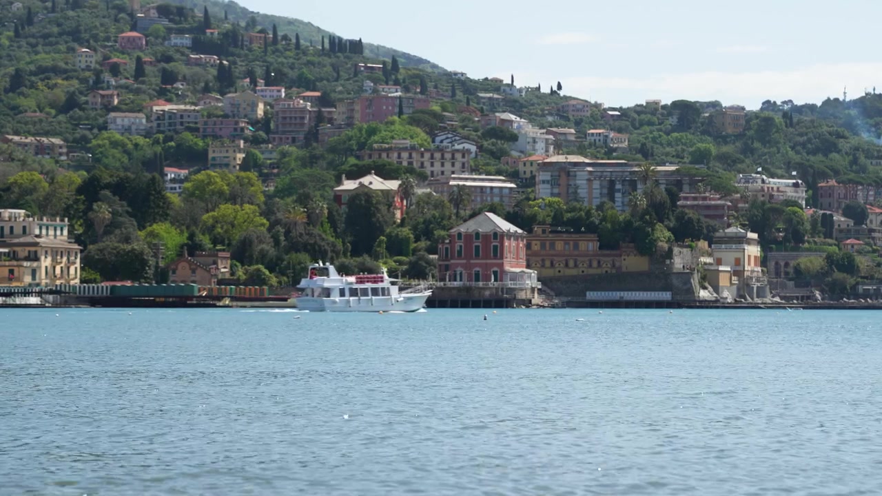 Inquadratura grandangolare degli edifici colorati sul lungomare di Rapallo, colline lussureggianti e una baia calma, mentre una barca turistica bianca scivola sull'acqua scintillante sotto la luce chiara del giorno. Nessuna persona visibile.