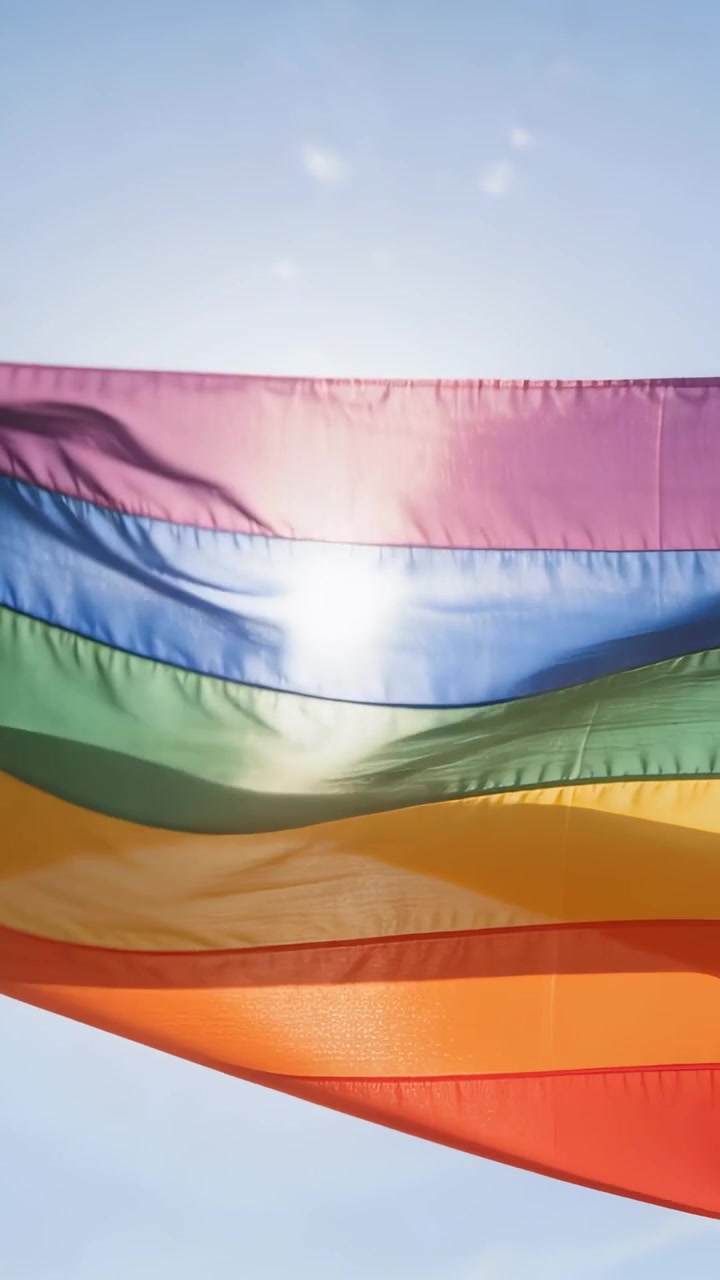 Video verticale: Grande bandiera arcobaleno che sventola nella brezza, mostrando le strisce contro il cielo blu, con il sole che splende.