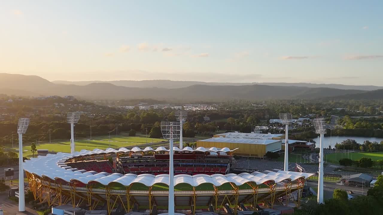 A high-angle drone shot captures a large circular sports stadium during a golden hour sunset