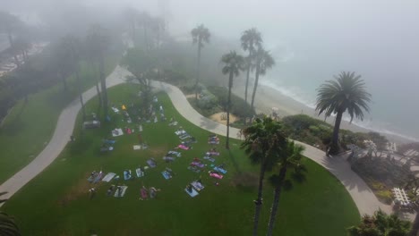 Vista-Aérea-De-Los-Pájaros-De-La-Gente-En-El-Césped-Del-Parque-Frente-A-La-Playa-Haciendo-Ejercicio-Matutino-Bajo-La-Niebla,-Disparo-De-Drones