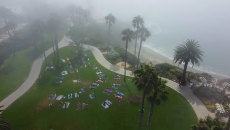 birds eye aerial view of people at beachfront park grass having morning exercise under mist, drone shot