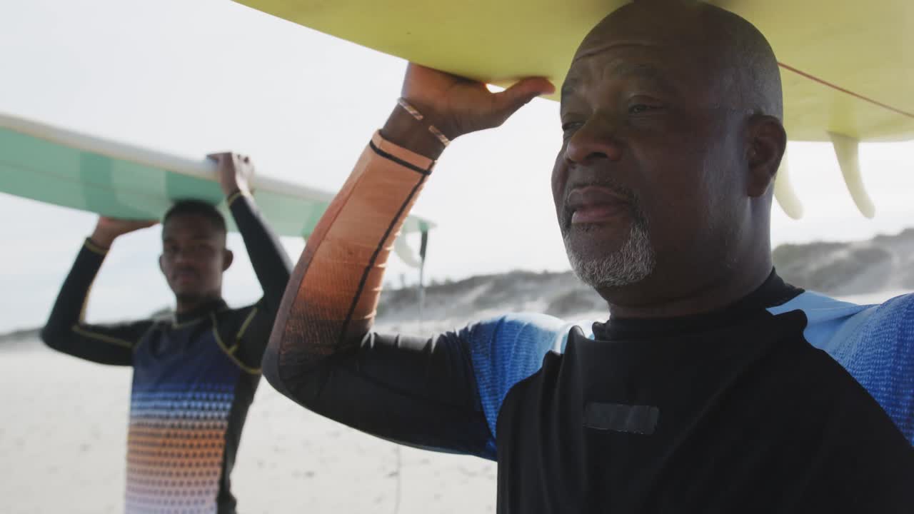 афроамериканский отец и сын-подросток стоят на пляже с досками для серфинга на головах и разговаривают