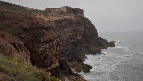 SLOW-MOTION-waves-white-with-foam-break-on-rocky-cliff-with-old-dark-castle-at-the-top,-Cape-Roca-Portugal