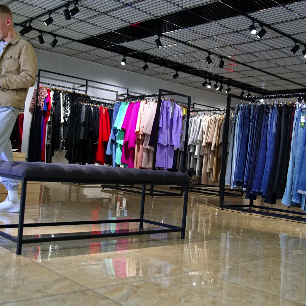 Young man with phone in hand walks by the clothes shop nervously. Male visitor expecting his girlfriend trying clothes in the fitting room
