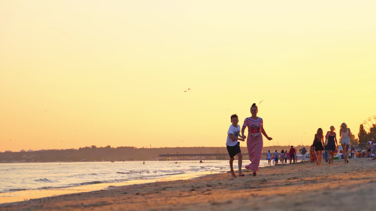 Joyful woman with a boy spending a good time at seaside. Happy mother is running together with her son on the seashore in the evening. Summer vacation.