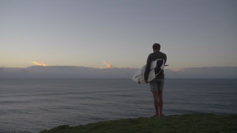 man standing on edge of cliff looking at ocean at sunrise holding surfboard