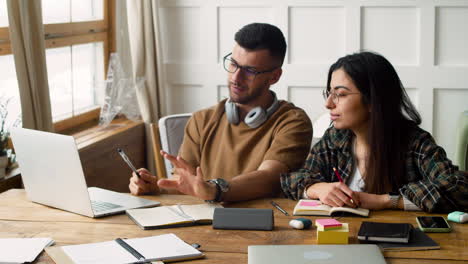 student with headphones talking with female mate using laptop on the table discussing about a project